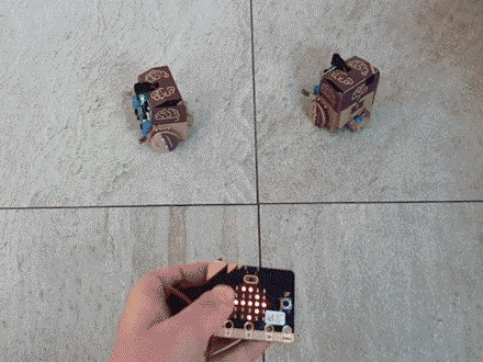 Looping animation of two small cube shaped robots moving in response to commands sent from a BBC micro:bit (a small circuit board with a matrix of LEDs on the front displaying different direction arrows)